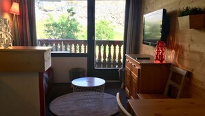 A cozy room with a wooden table, a round white table with a wireframe base, a TV on the wall, and a window with a view of greenery outside. - T2-6pers-34m2-quartier Bergers-pied des Pistes (Huez)