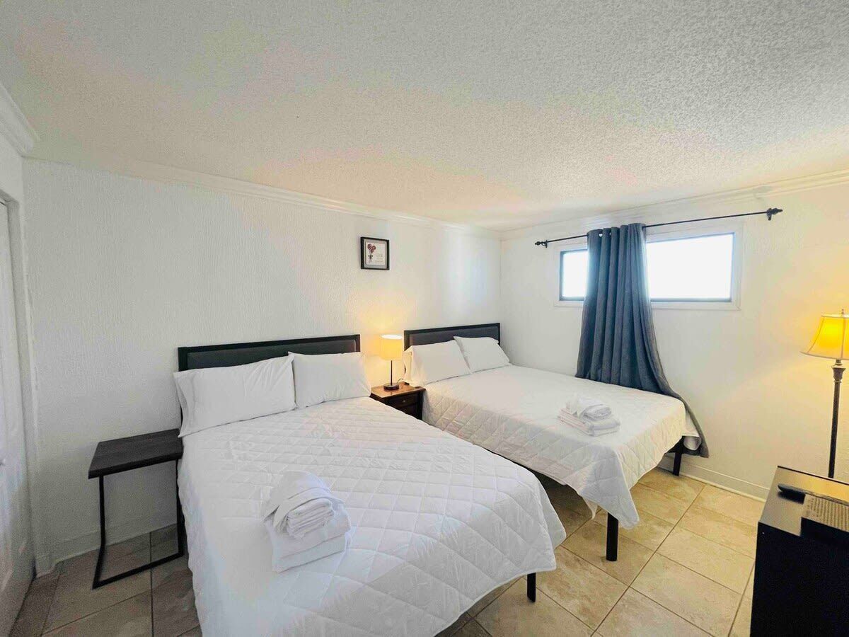 Une chambre d’hôtel avec deux lits, une petite table, une fenêtre avec des rideaux et une lampe.