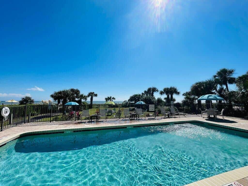 Una piscina azul cristalina con una solana empedrada, rodeada de palmeras y sombrillas, bajo un cielo soleado.