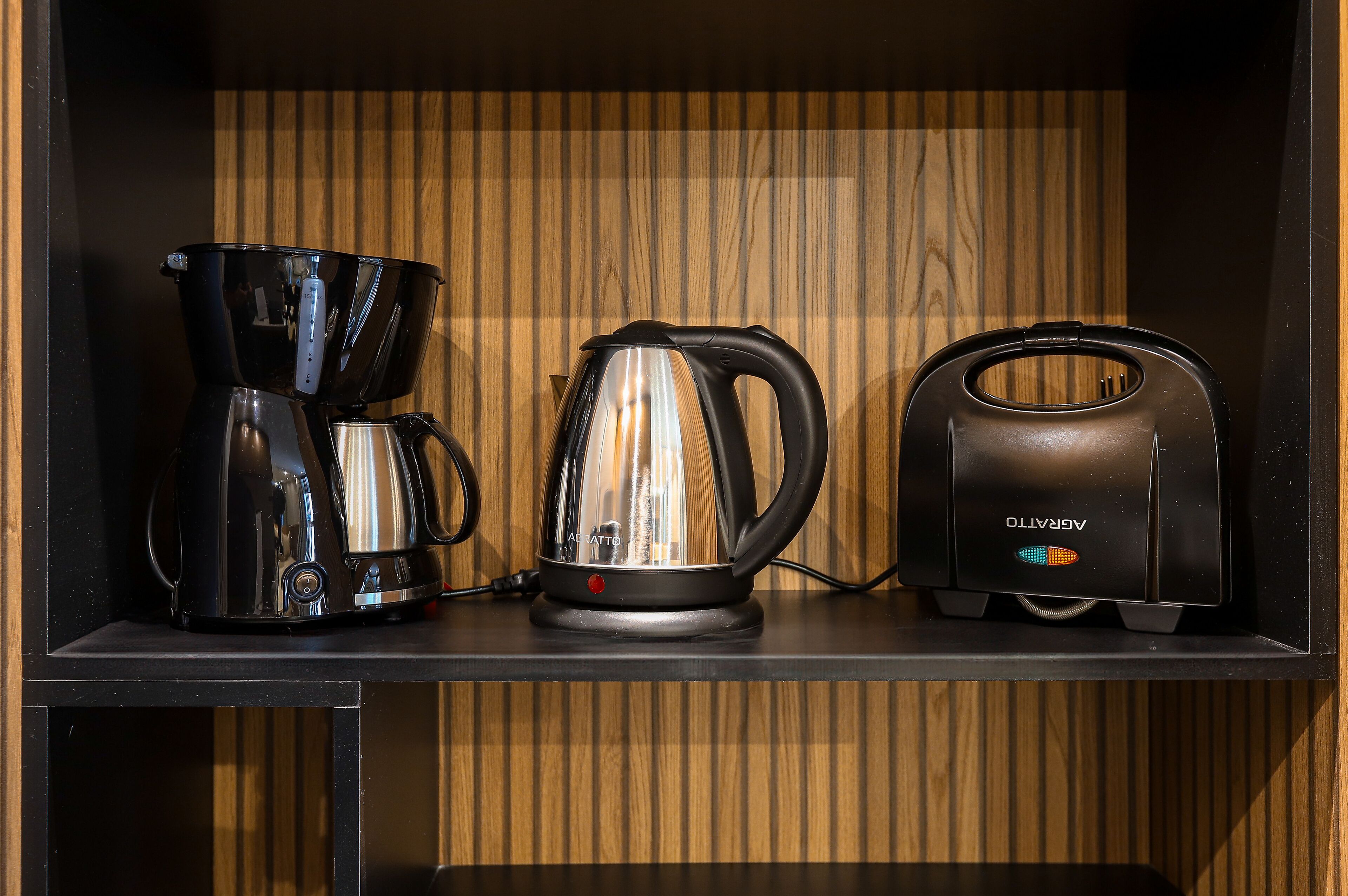A kitchen area with a coffee maker, kettle, and toaster on a black countertop against a wooden wall.