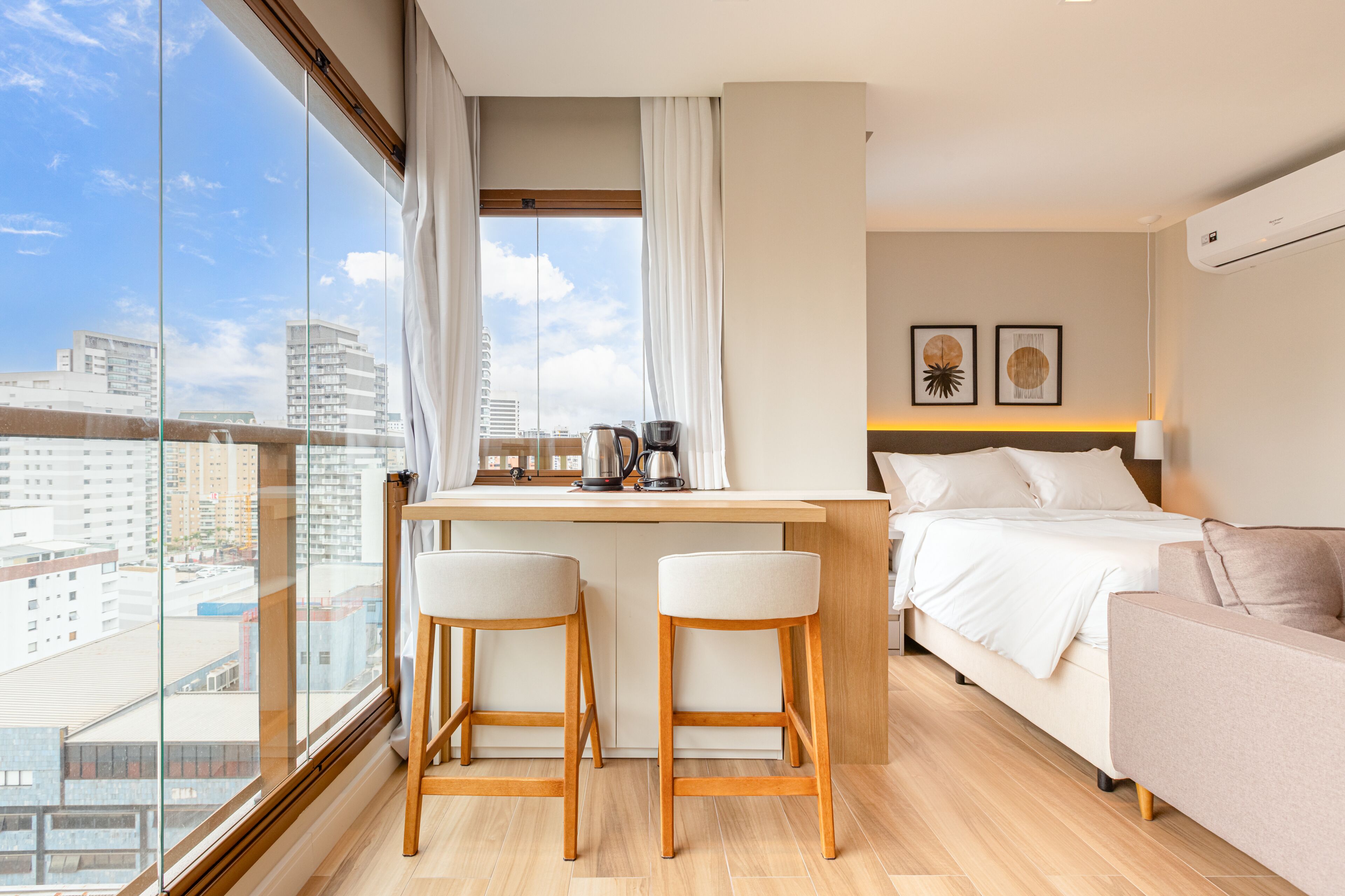 A modern hotel room with a large window, a bed, a sofa, and a small kitchen area.