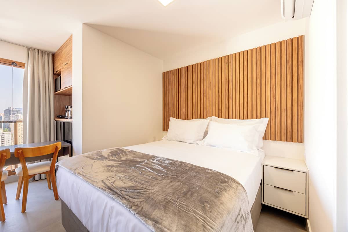 Ein Hotelzimmer mit einem grossen Bett, einem Holzkopfstück, einem Schreibtisch mit Stuhl und Blick auf die Stadt durch das Fenster.