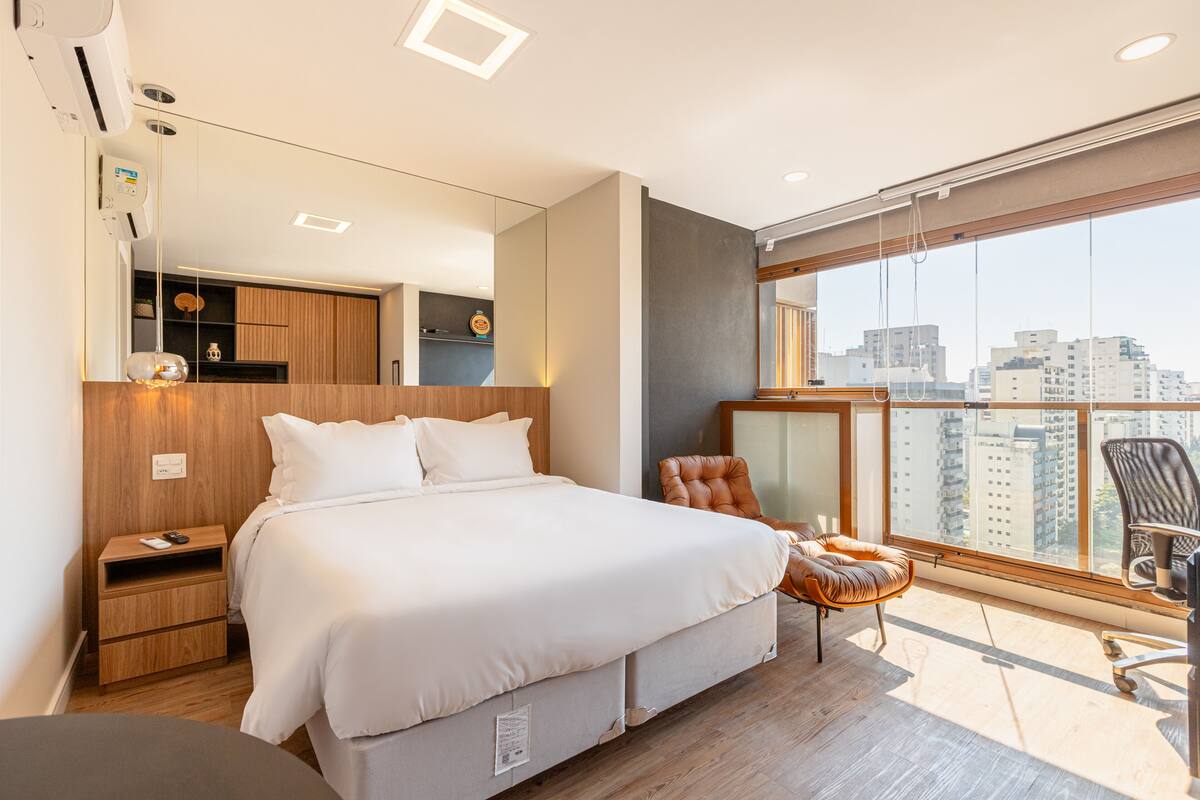 Ein modernes Hotelzimmer mit einem großen Bett, einem Holfnachtkästchen, einem Sessel und einem Balkon mit Blick auf die Stadt.