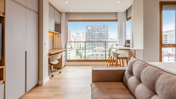 A modern apartment with a large window, a sofa, a desk, and a chair. - DVN - D'Oru Vila Nova Conceição by Anora (São Paulo)