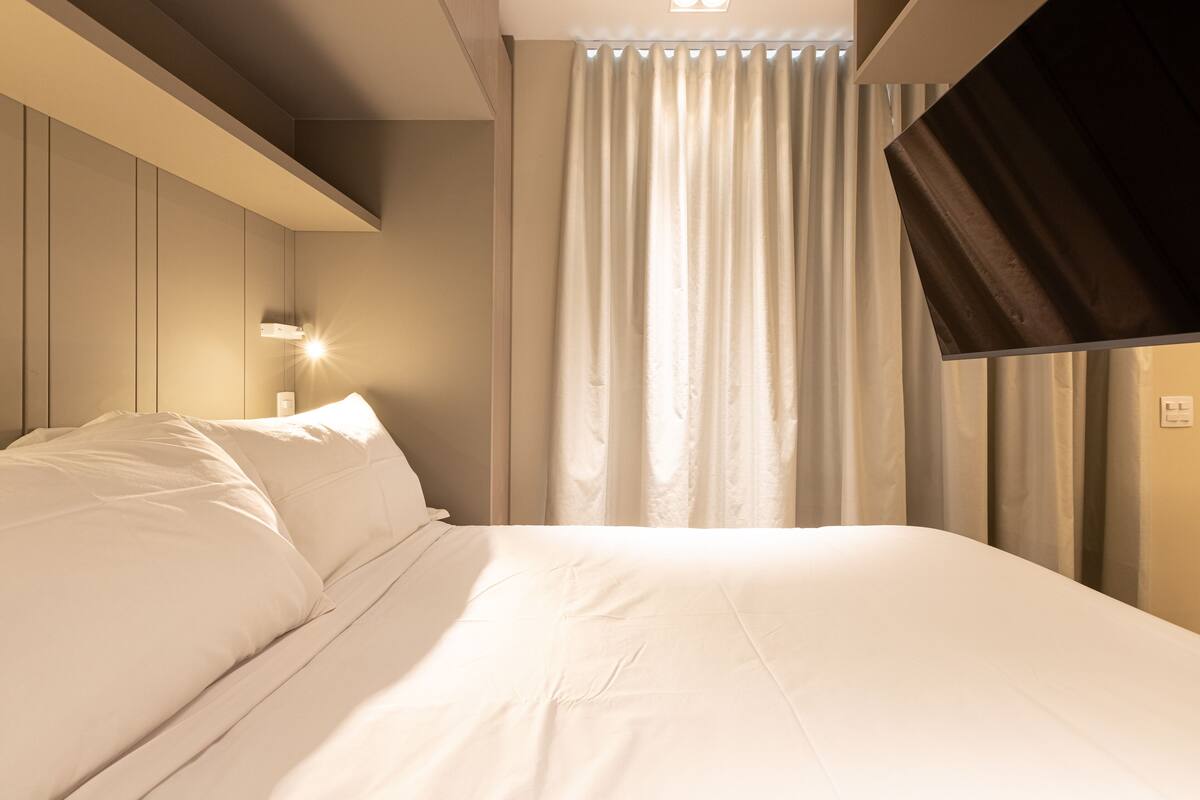 Ein ordentlich bezogenes Bett in einem Hotelzimmer mit Kopfteil, Wandleuchte und einem Vorhangfenster.