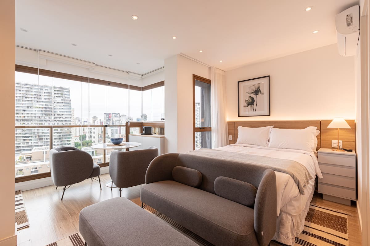 Ein modernes Hotelzimmer mit einem großen Bett, einem Schreibtisch, zwei Stühlen und Blick auf die Stadt durch große Fenster.