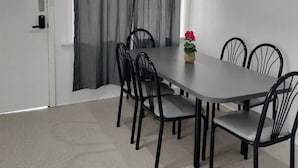 Un coin repas avec une table rectangulaire, six chaises noires, des rideaux gris et une plante en pot ornée de fleurs rouges.
