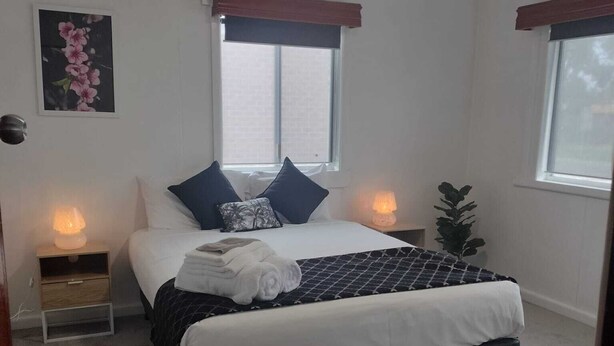 Une chambre à coucher comprenant un lit, deux tables de chevet, une commode, une fenêtre avec des rideaux, un luminaire fixé au mur et une plante en pot.