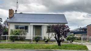 Une maison de plain-pied avec un toit gris et un revêtement extérieur blanc, une petite cour à l’avant avec un arbre, et une clôture en métal.