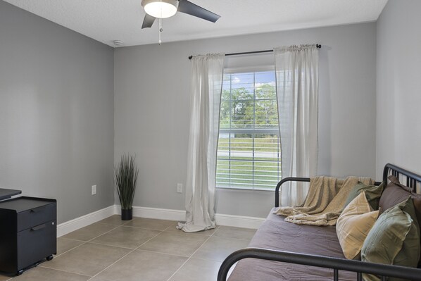 A bedroom with a bed, a ceiling fan, a window with curtains, a potted plant, and a desk.