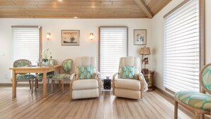 A spacious living room with wooden flooring, a dining table with chairs, and large windows with blinds. - Beachside Cottage
 (Flagler Beach)