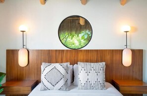 Un dormitorio con cabecera de madera, dos mesitas de noche, un espejo redondo y vistas a la naturaleza, junto con almohadas estampadas.