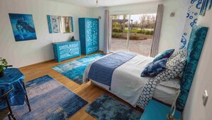 A bedroom with a bed, a dresser, a desk, and a large window with a view of the outdoors. - Chez Phil, Chambres D'hôte, Chambre Thème Eau (La Pomarède)