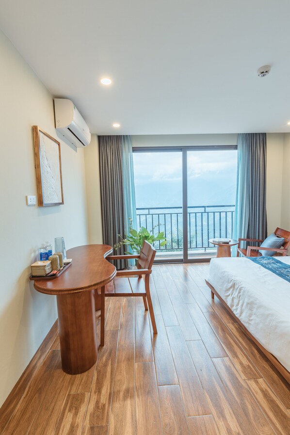A hotel room with a wooden floor, a bed, a desk with a chair, a small table with a plant, and a balcony with a view.