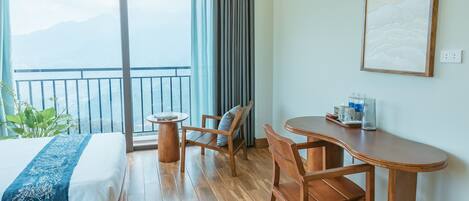 A hotel room with a bed, a desk, two chairs, a small table, a window with a view of mountains, and a painting on the wall.