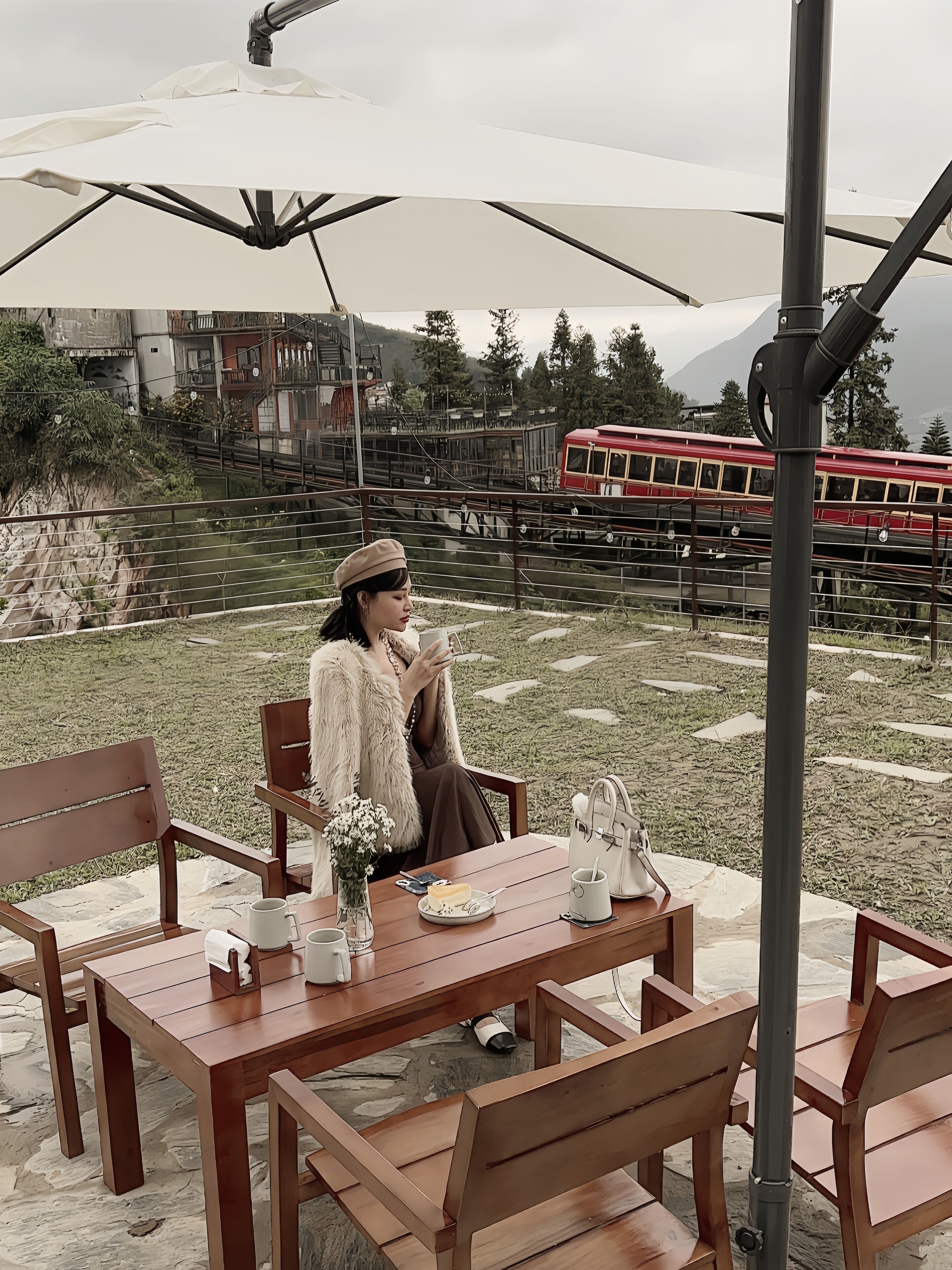 Una persona sentada en una mesa al aire libre con una sombrilla blanca, un carrito rojo al fondo y un paisaje montañoso.