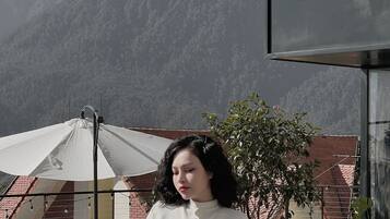 Eine Person steht im Freien vor einer Bergkulisse, mit einem weißen Schirm und einem Gebäude mit rotem Dach.