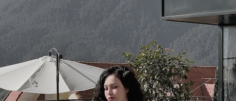 Una persona está de pie al aire libre, con montañas al fondo, un paraguas blanco y un edificio de techo rojo.