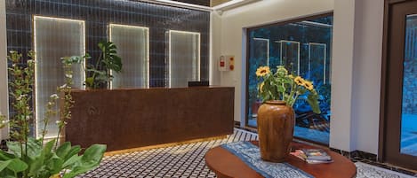 A lobby with a reception desk, patterned floor tiles, a large vase with flowers, and a potted plant.