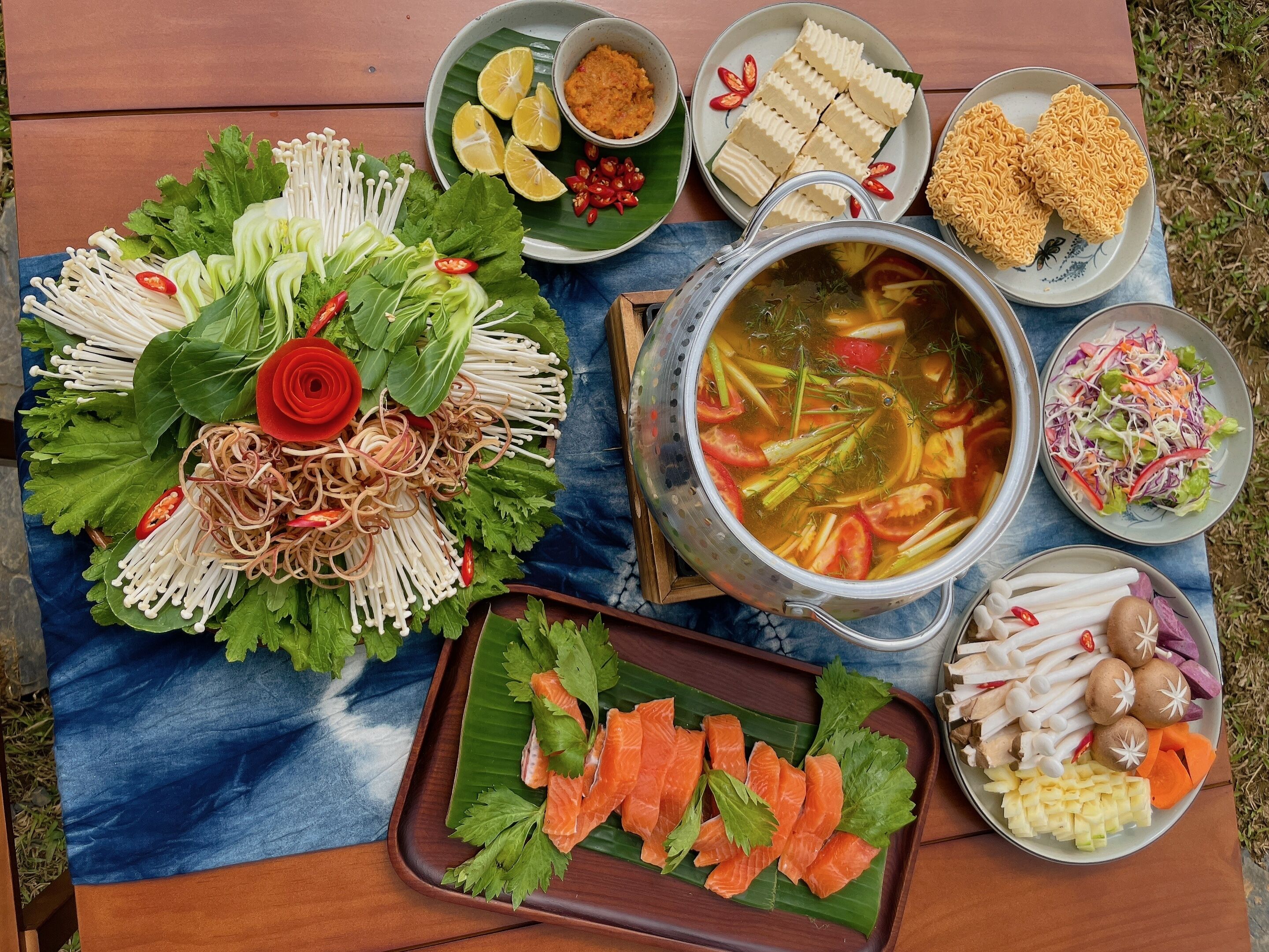 A variety of dishes including a pot of soup, fresh vegetables, and sliced meat, arranged on a wooden table outdoors.