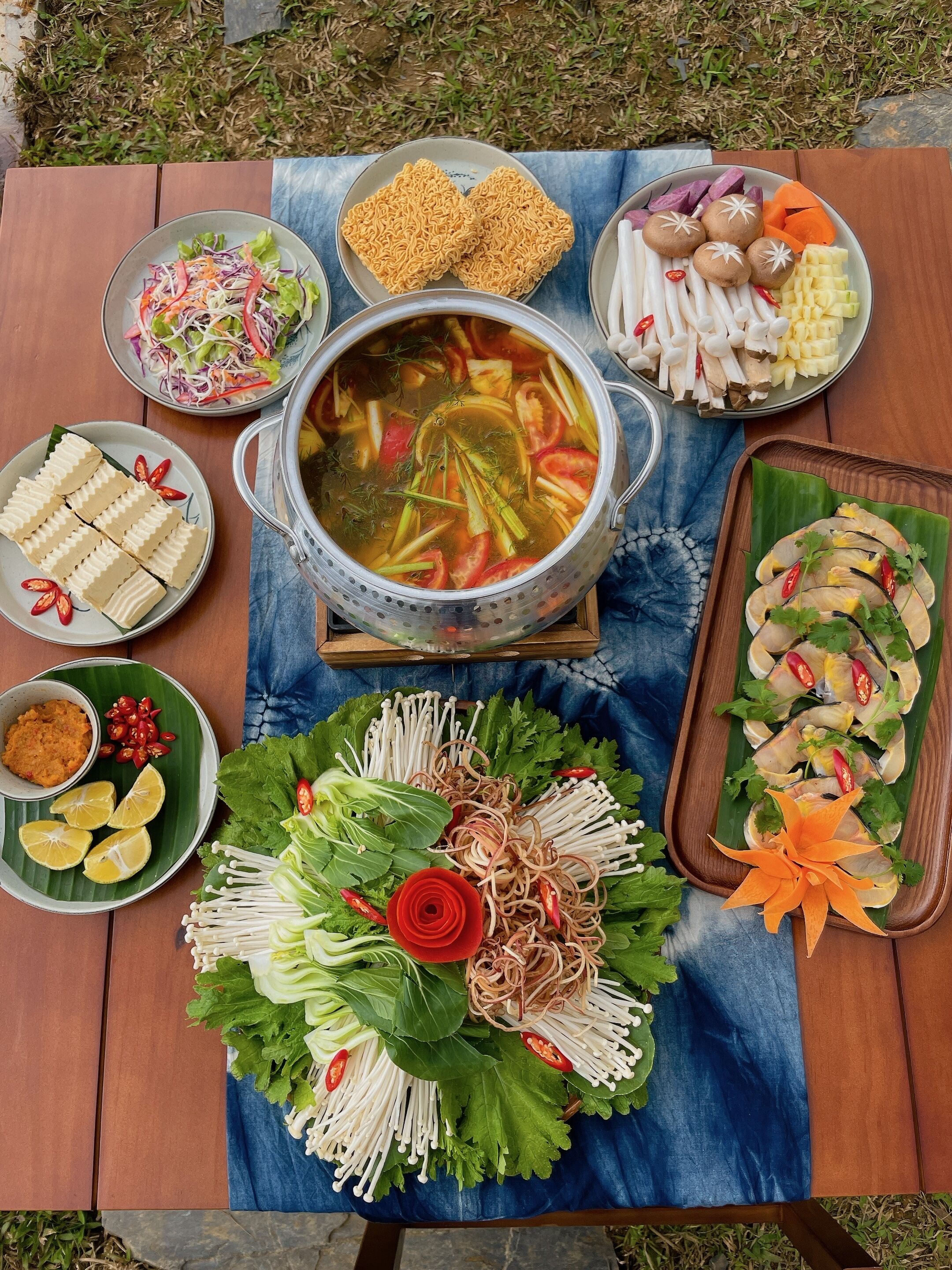 A table with various dishes including a pot of soup, plates of noodles, and fresh vegetables.