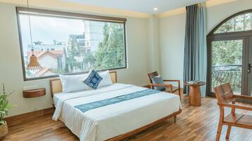 A bedroom with a large bed, wooden flooring, a window with a view of buildings, a balcony with a chair, and a small table.