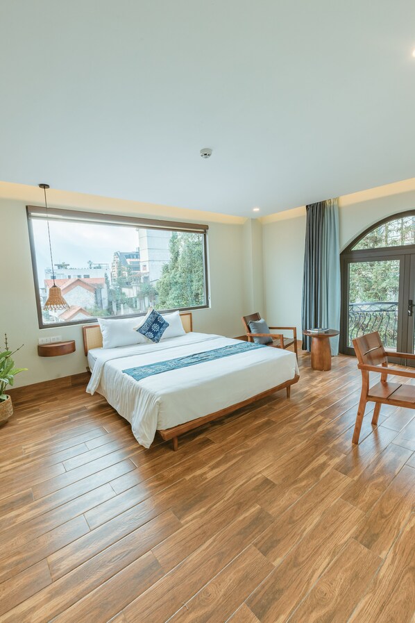 A bedroom with a large bed, wooden flooring, a window with a view of buildings, a balcony with a chair, and a small table. - Sapa YEN Hotel (Sa Pa)