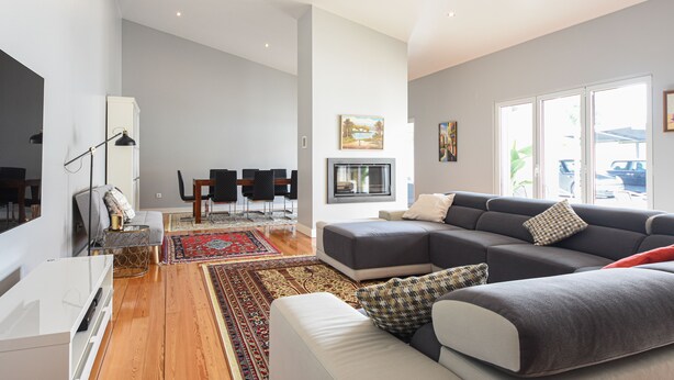 A modern living room with a grey sectional sofa, a white ottoman, and a red throw pillow. There is a dining area with a table and chairs, and a large window providing natural light. - Casa dos Infantes  (Vendas Novas)