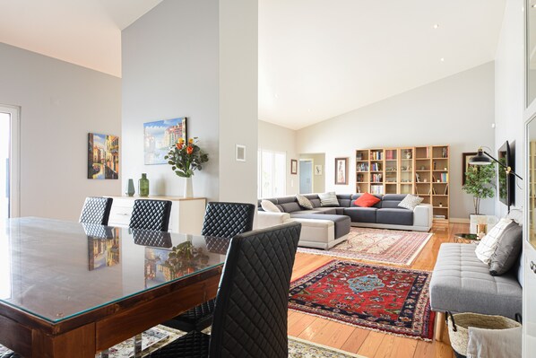 A spacious living room with a large glass dining table, comfortable seating, a bookshelf, and a red patterned rug.