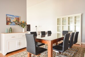 A dining area with a glass-top table, black cushioned chairs, a white cabinet, and a painting of a canal scene.