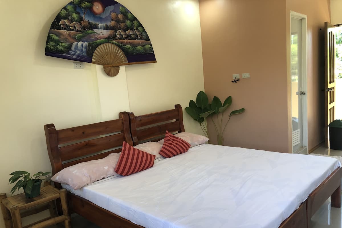 Ein Schlafzimmer mit einem hölzernen Bett, weißer Bettwäsche, einem Nachttisch mit einer Pflanze und einem Wandventilator mit Landschaftsbild.