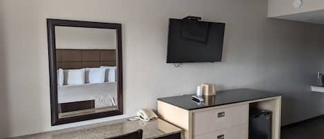 Habitación de hotel con cama, escritorio con silla, televisión, minibar y espejo.