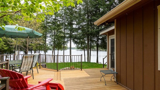 Una terraza de madera con mobiliario exterior, vista a un lago y un edificio con una puerta.