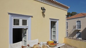 A table set with food and drinks on a balcony with white chairs, a yellow building with white trim, and a tiled roof.