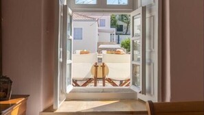 A room with a window opening to a balcony, a chair with a cushion, and a small table with a chair.
