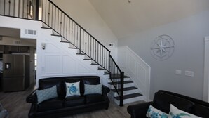 Un amplio salón con un sofá de cuero negro, una escalera con barandillas negras y una puerta blanca.