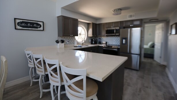 Una cocina moderna con una isla grande, sillas blancas y un letrero que dice 'Gather Here Always'.