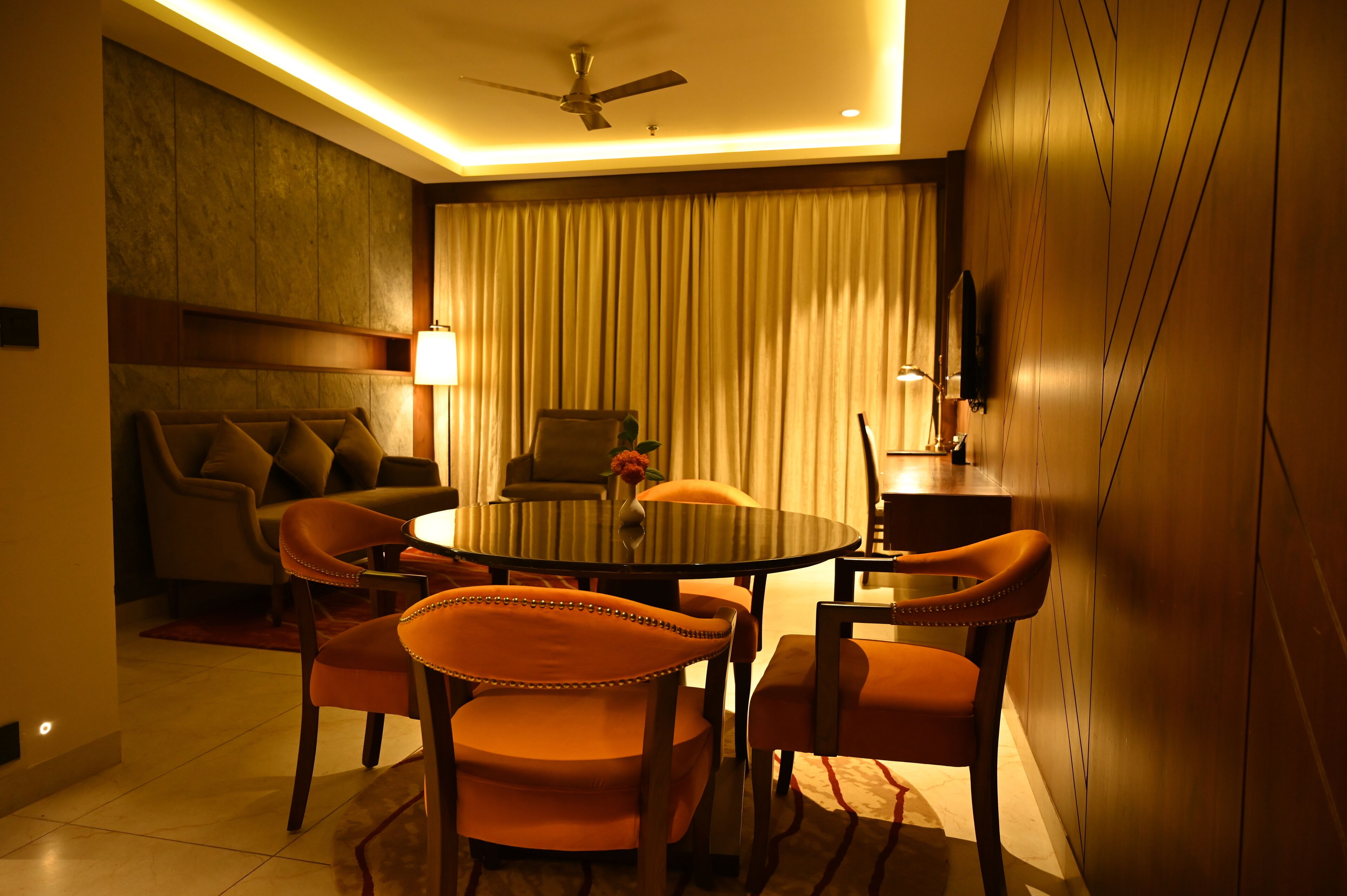 A modern hotel room with a round dining table, four orange chairs, a sofa, a ceiling fan, and a large window with curtains.