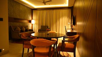 A modern hotel room with a round dining table, four orange chairs, a sofa, a ceiling fan, and a large window with curtains.