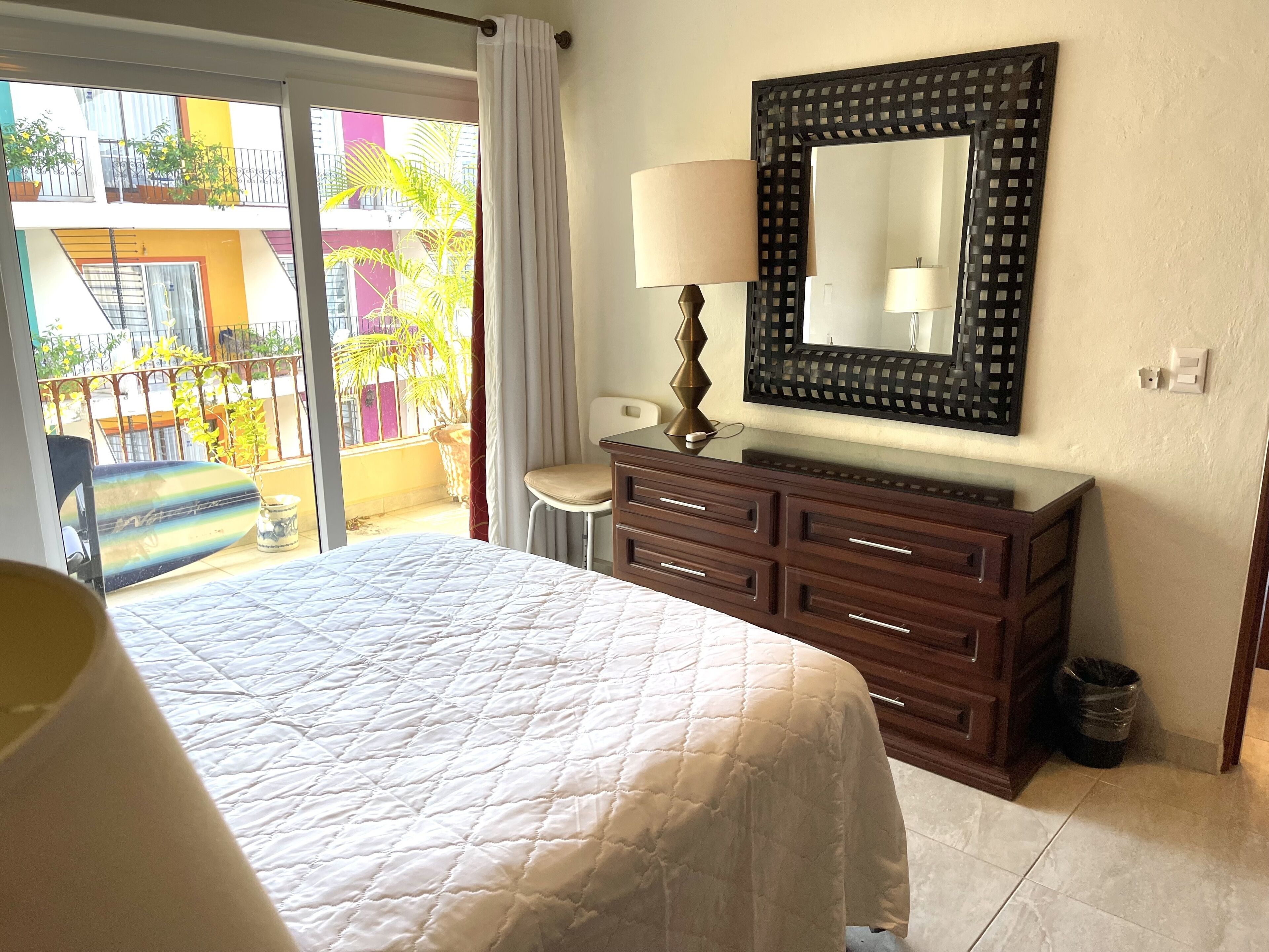 Une chambre d’hôtel avec un lit, une commode, un miroir et une vue sur un balcon agrémenté de plantes et donnant sur un immeuble.