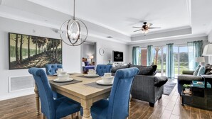 Ein modernes Wohnzimmer mit Essbereich, mit einem großen Gemälde, blauen Polsterstühlen, einem dunklen Sofa und einem Deckenventilator.