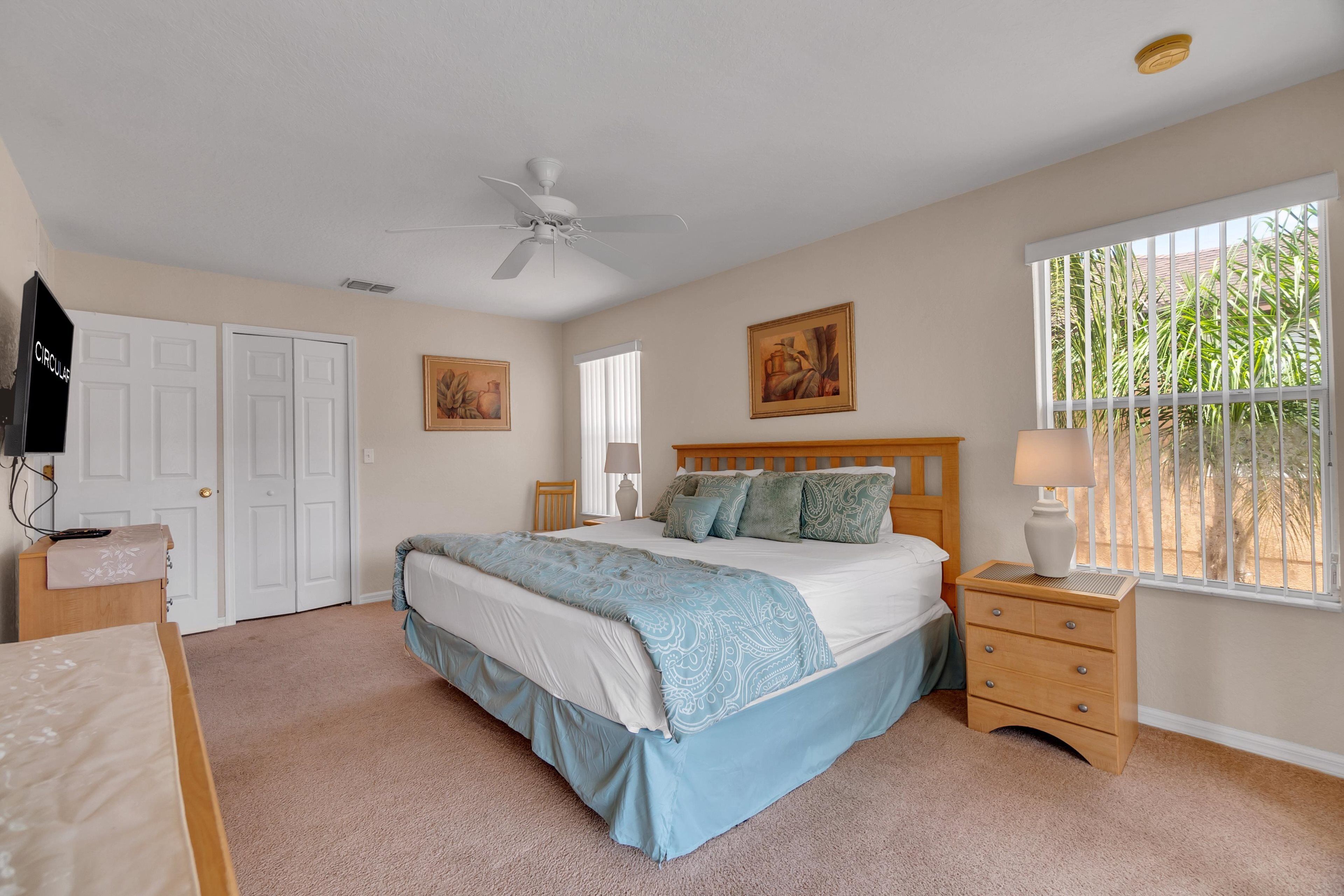 A bedroom with a bed, a nightstand, a ceiling fan, a window with a view of palm trees, and a door.