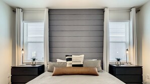 Ein Schlafzimmer mit einem großen Bett, zwei Nachttischen mit Lampen, einem dunklen Kopfteil und einem Fenster mit Jalousien.