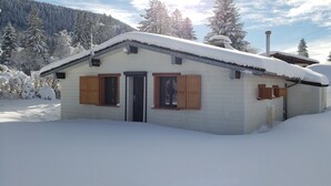 Una pequeña casa de un solo piso con techo inclinado cubierto de nieve, rodeada de un paisaje nevado y árboles.