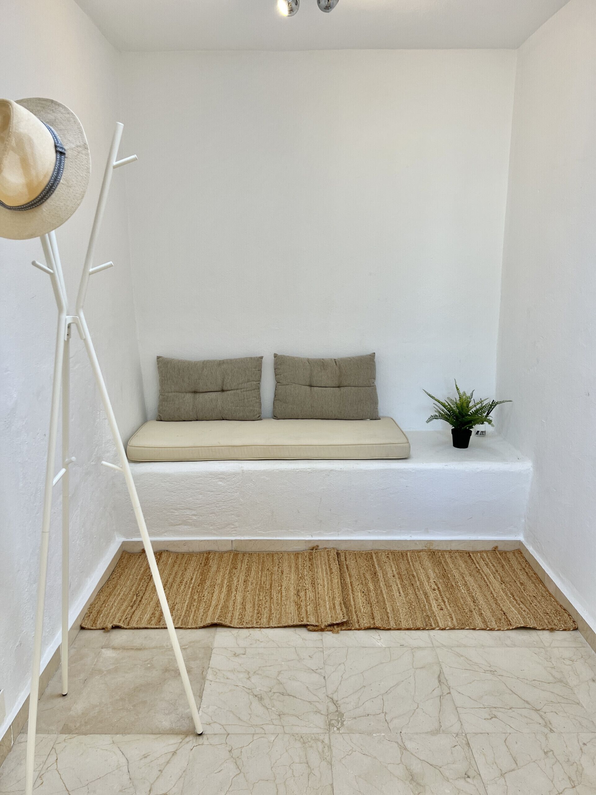 Ein Eckzimmer mit einer weißen Bank, zwei Kissen, einer Stehlampe, einem kleinen Topfpflanzen und einem strukturierten Teppich.