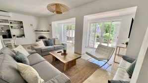 A modern living room with a grey sofa, a wooden coffee table, and a white armchair. There is a fireplace, a dining area, and a view of the outdoors through large windows. - 58 -Villa, private swimming pool, close to the sea (Estepona)