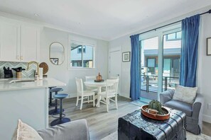 A modern kitchen with white cabinets, a dining area with a round table and chairs, a bar area with stools, and a living room with a sofa and a coffee table.