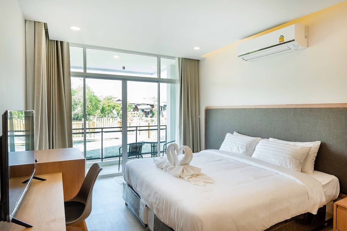 Habitación de hotel con cama, escritorio y silla. Se ve un ventanal con cortinas, y un balcón con mobiliario exterior a través de la ventana.