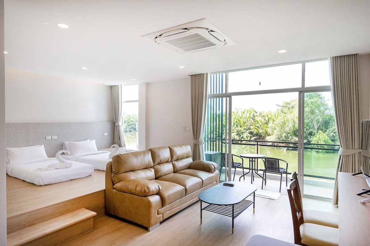 Habitación de hotel moderna con sofá, cama, balcón con mesa y sillas, y un ventanal con vistas al verdor.