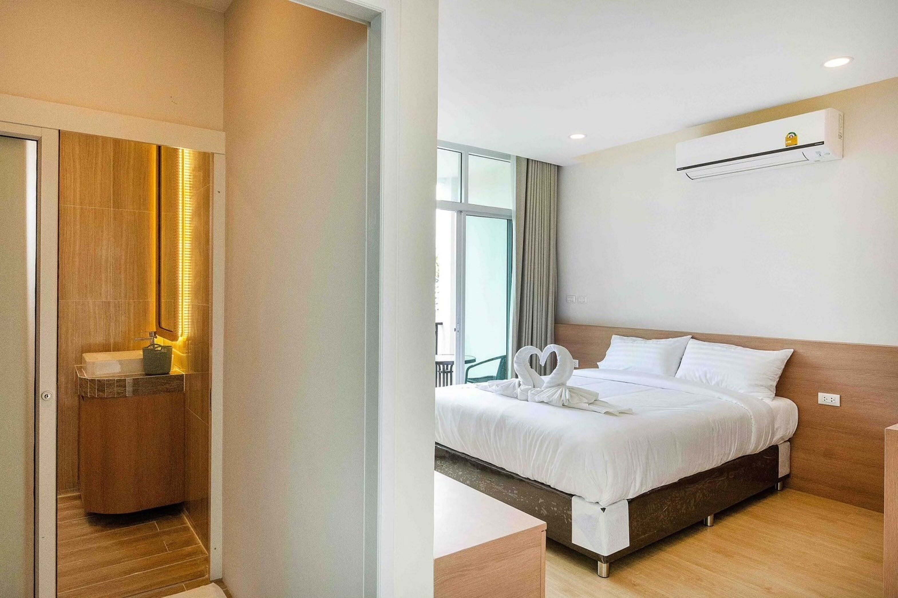 A hotel room with a wooden vanity, a bed with white bedding, a wall-mounted air conditioner, and a large window with curtains.